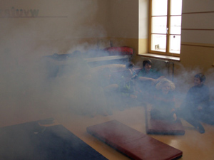 Das Foto zeigt einen KLassneraum mit einigen Schülern; durch den Raum ziehen dichte Nebelschwaden.