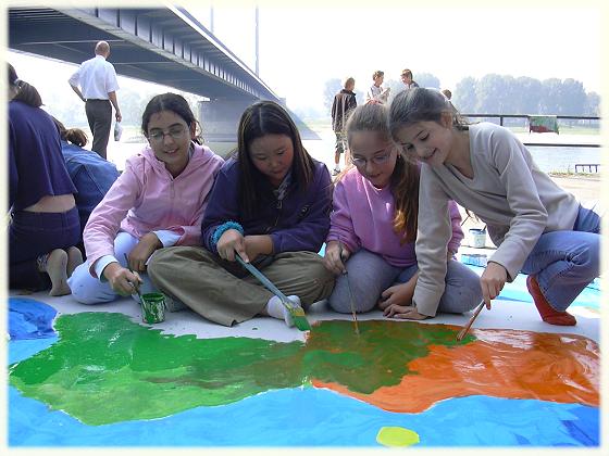 Das Foto zeigt vier Mädchen, die am Rheinufer auf einer großen Stoffbahn malen.