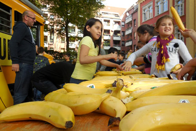 Das Foto zeigt einen Tisch voller Bananen. Im Hintergrund sind Kinder, ein Mann und ein gelber Truck zu sehen.