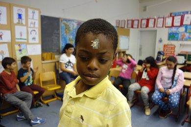 Das Foto zeigt einen Jungen in einem Klassenraum; er hat einen weißen Fleck auf die Stirn gemalt. Im Hintergrund sitzen weitere Schüler.