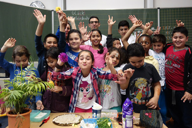 Das Foto zeigt Kinder verschiedener Nationalität, die in ihrem KLassenraum vor der Tafel stehen und begeistert winken.
