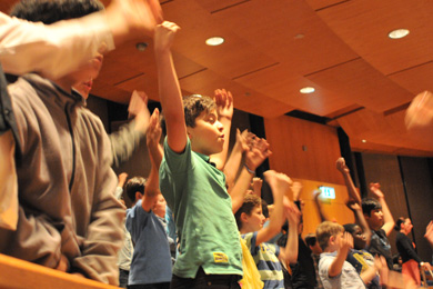 Das Foto zeigt viele singende Kinder in einer Konzerthalle.