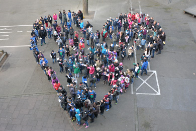 Das Foto zeigt Schüler und Lehrer, die auf dem Schulhof stehen und gemeinsam ein großes Herz bilden.