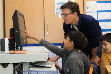 Das Foto zeigt zwei Jungen, die vor einem Computer sitzen und ihrem Lehrer auf dem Bildschirm etwas zeigen.