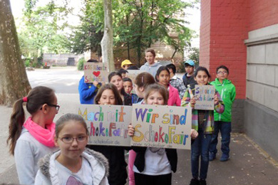 Das Foto zeigt Kinder, die vor ihrer Schule laufen und Plakate zur Schokofair-Aktion inden Händen halten.