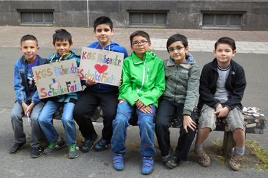Das Foto zeigt Kinder, die auf einer Bank auf ihrem Schulhof sitzen und Plakate zur Schokofair-Aktion inden Händen halten.