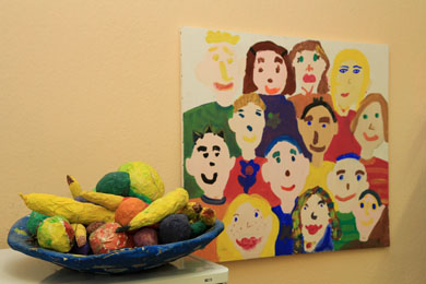 Das Foto zeigt im Vordergrund eine Schale mit aus Pappmaschee gefertigtem Obst und im Hintergrund ein farbenfrohes gemaltes Bild, das eine Gruppe von freundlichen Menschen darstellt.