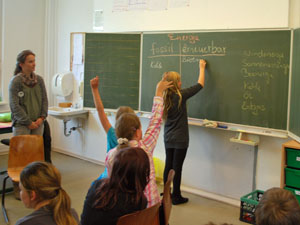 Das Foto zeigt einen Klassenraum mit Schülerinnen und Schülern. Eine Schülerin steht an der Tafel und schreibt in eine Tabelle, in der fossile und erneuerbare Energieformen aufgelistet sind.