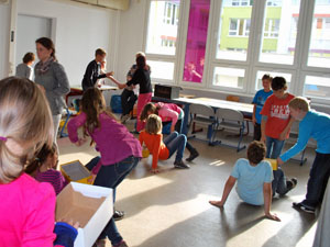 Das Foto zeigt Schülerinnen und Schüler, die in einem Klassenraum sehr eifrig kleine Pappkartons durch die Gegend tragen.