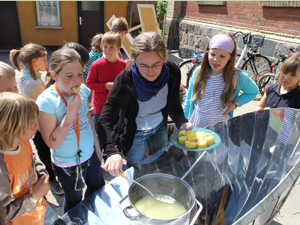 Das Foto zeigt eine Frau, die gedünstete Maisscheiben aus einem großen Topf herausfischt. Dieser Topf wird auf einem Solarkocher - einer Parabolschüssel aus poliertem Metall - erhitzt. Eine Gruppe von Kindern steht darum und freut sich schon auf den leckeren Mais.