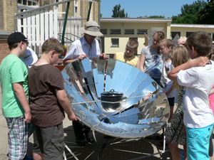 Das Foto zeigt Schüler, die im einen Solarkocher herum stehen. Ein Mann mit Sonnenhut erklrt ihnen den Kocher.