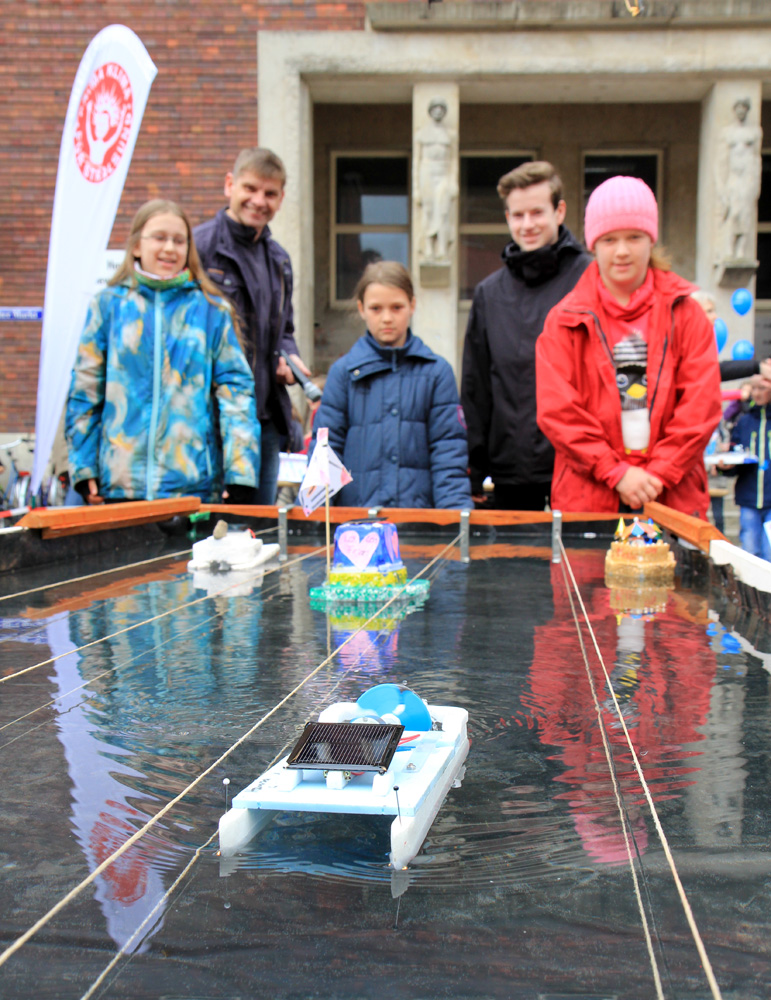 Das Foto zeigt Solarboote auf einer Rennstrecke; im Hintergrund die Schüler, die diese Boote gebaut haben.