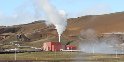 Das Foto zeigt die Geothermalregion Krafla auf Island. Es ist eine karge Graslandschaft zu sehen, die von Rohrleitungen durchzogen wird. In der Bildmitte steht ein Kraftwerk; es steigt weißer Dampf auf.