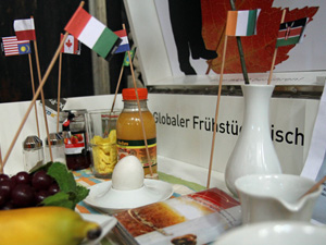 Das Foto zeigt verschiedene Nahrungsmittel - eine Banane, ein Ei, eine Flasche Fruchtsaft etc.; diese sind jeweils mit kleinen Flaggen versehen, welche das Herkunftsland zeigen.