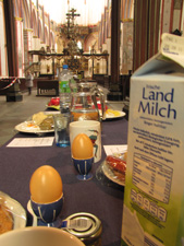 Das Foto zeigt im Vordergrund eine mit mehreren verschiedenen Mahlzeiten gedeckte Tafel, neben jeder Mahlzeit liegt die CO2-Rechnung. Den Hintergrund bildet das Hauptschiff der Nikolaikirche Stralsund, wo die Installation im Oktober 2011 zu sehen war.