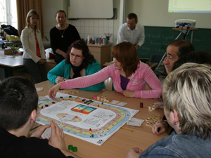 Schüler und Lehrer sitzen an einem Tisch und spielen das Simulationsspiel "TriCO2lor"; im Hintergrund weitere Lehrer sowie der Spielleiter