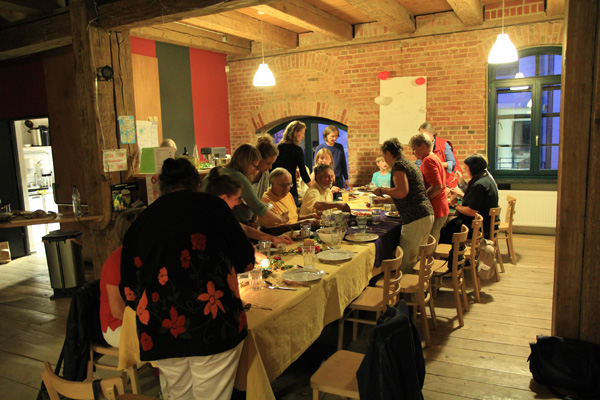 Das Foto zeigt einen großen Raum mit einer großen Tafel, an der Menschen sitzen bzw. stehen und sich Speisen auf ihre Teller tun.