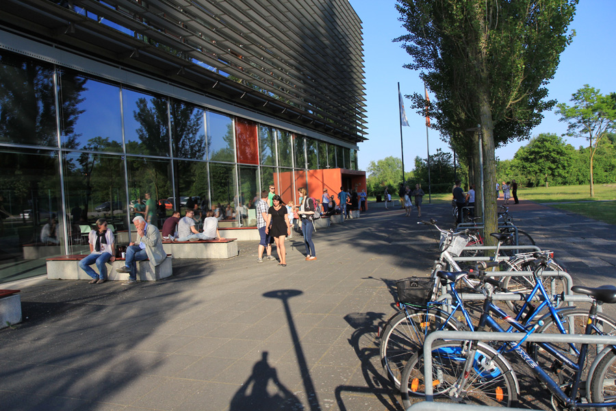 Das Foto zeigt den Block 7 der Hochschule Wismar von außen bei sommerlichem Licht. Davor stehen Fahrräder und sitzen Menschen.