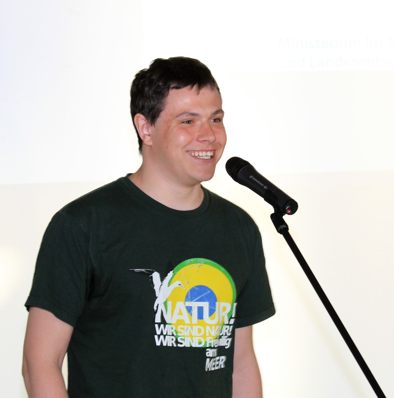Das Foto zeigt das Brustbild eines jungen Mannes, der in ein Mikrofon spricht.