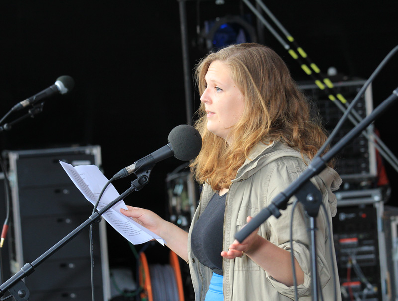 Das Foto zeigt eine Frau, die auf einer Bühne steht und in ein Mikrofon spricht.