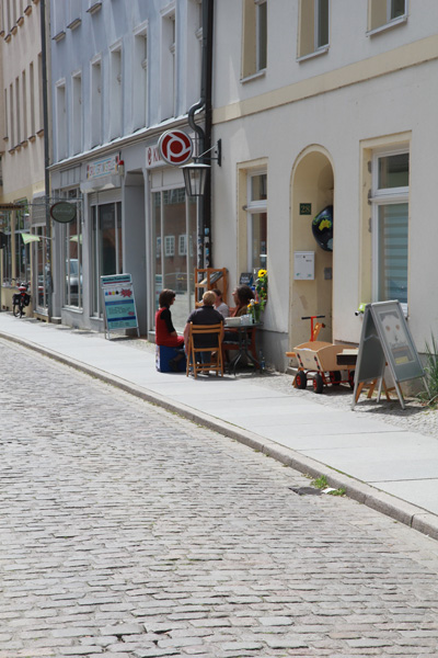 Das Foto eine ruhige Straße; auf dem Bürgersteig vor einem Haus sitzen vier Menschen auf einer Bank und Stühen, sie unterhalten sich..