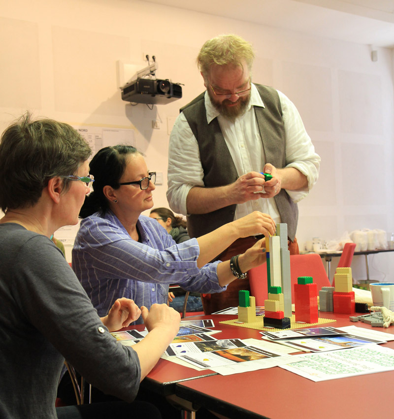 Das Foto zeigt drei Menschen in einem Seminarraum, die mit Papier und Legosteinen hantieren.