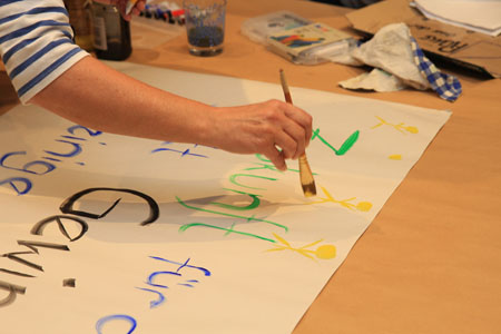 Ein Transparent wird gemalt. Das Foto zeigt eine Hand mit einem Pinsel sowie ein Blatt Papier mit der Aufschrift Zukunft für alle statt Gewinn für einige